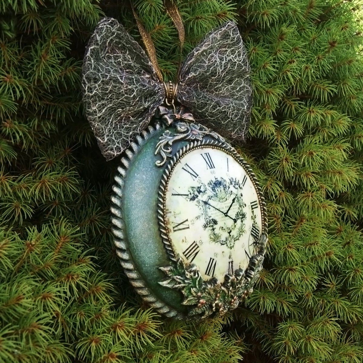 A decorative hanging ornament shaped like an antique clock, featuring a distressed teal-green oval body, white clock face with black Roman numerals, lace bow at the top, and leafy embellishments, photographed against green pine branches.
