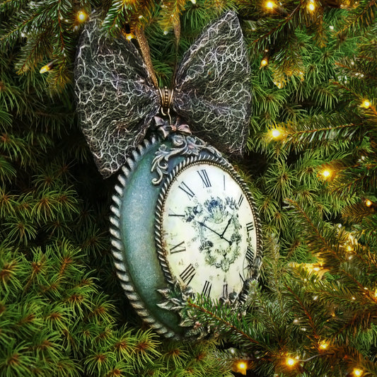 n antique-style decorative medallion- clock with a large textured gray bow on top, featuring a distressed green frame, Roman numerals, and a floral crest on the clock face.