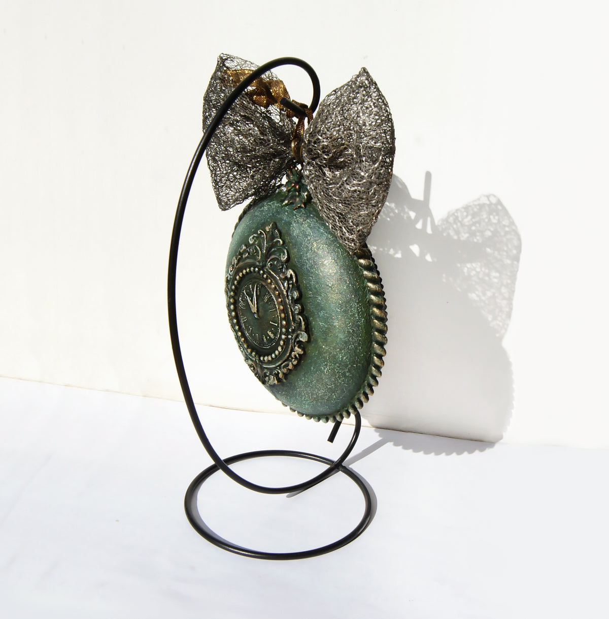 A decorative Christmas ball-clock with a distressed teal-green face and ornate silver-toned metal detailing, displayed on a black wire stand against a white background.