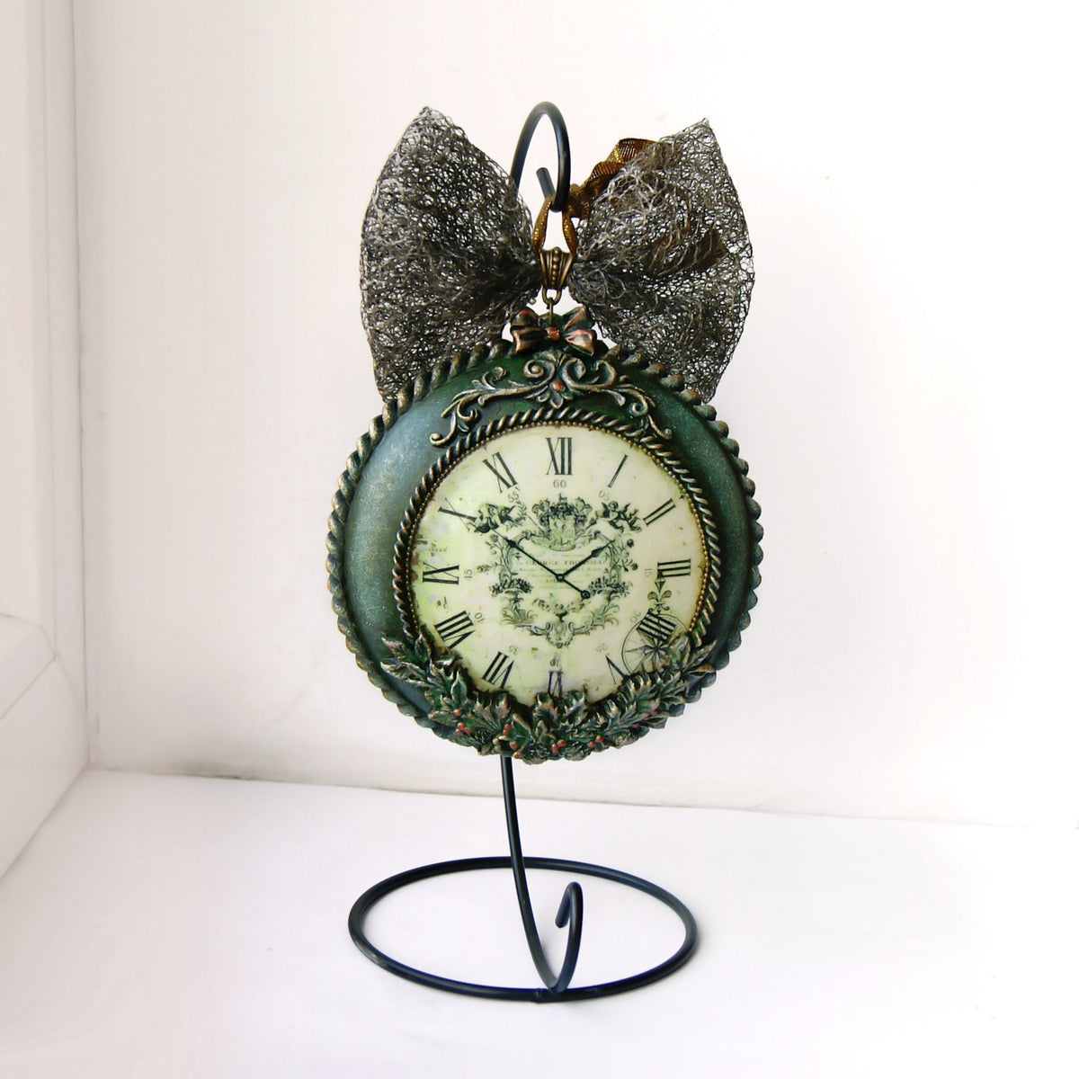 A decorative Christmas ball-clock with a distressed teal-green face and ornate silver-toned metal detailing, displayed on a black wire stand against a white background.