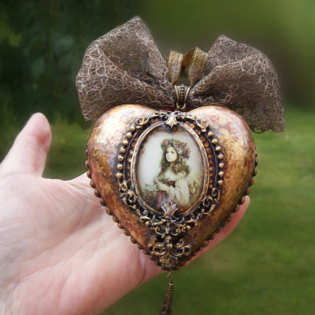An antique Victorian Christmas ball shaped like a heart, featuring a central oval portrait of a girl, ornate filigree detailing, a textured metallic bow at the top, and a long black tassel hanging from the bottom.