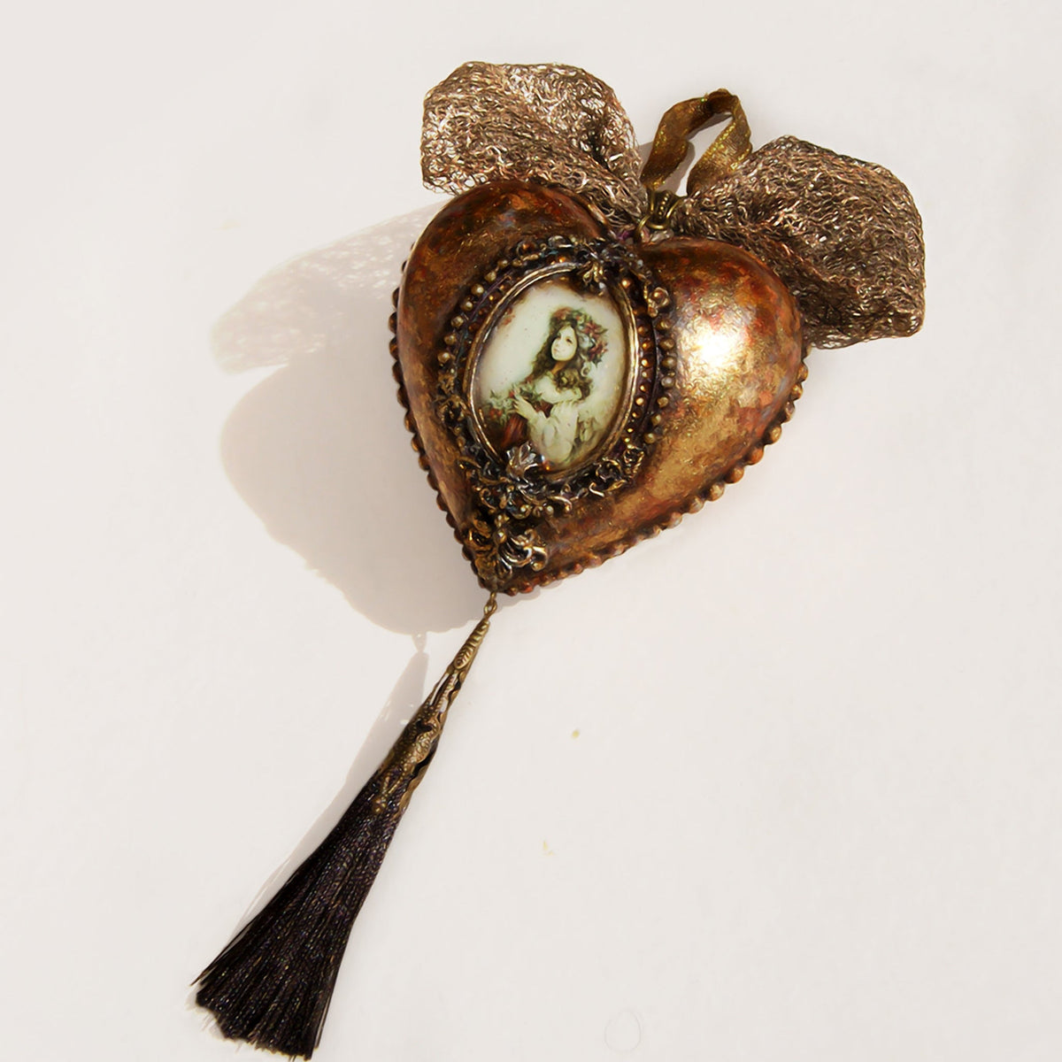 An antique Victorian Christmas ball shaped like a heart, featuring a central oval portrait of a girl, ornate filigree detailing, a textured metallic bow at the top, and a long black tassel hanging from the bottom.